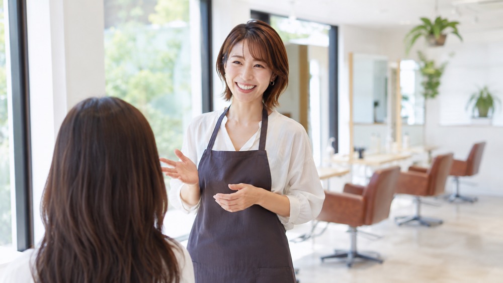 美容室でエプロン姿の女性美容師が笑顔で話しかけている様子