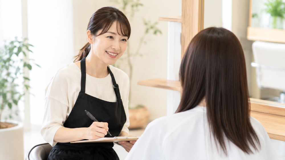 美容室でエプロン姿の女性美容師が笑顔で話しかけている様子