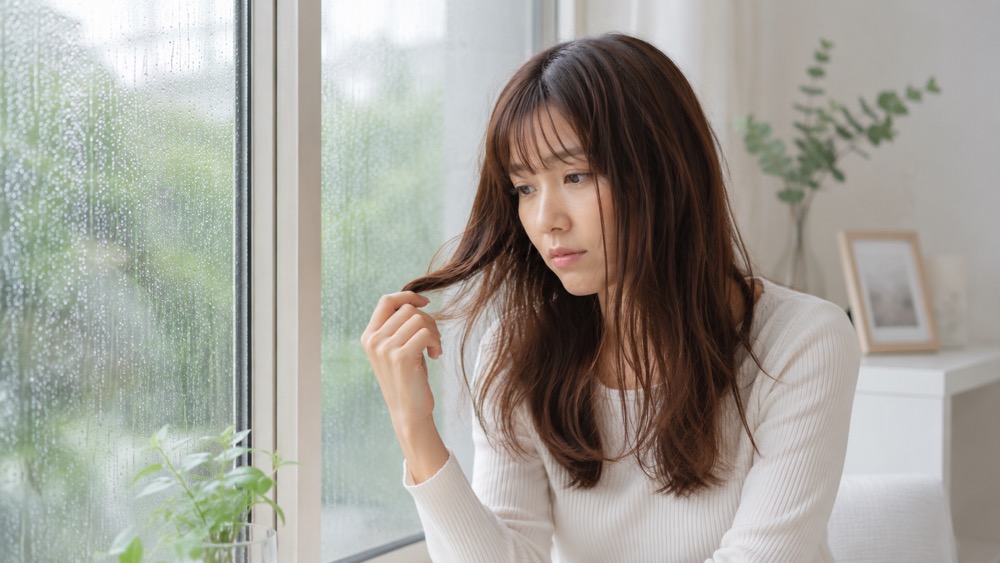 梅雨の窓辺で髪のうねりを気にする女性
